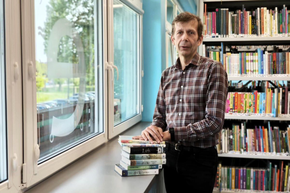 Mężczyzna w brązowej koszuli w kratę stoi w bibliotece przy oknie z widokiem na zieleń, oparty o parapet, na którym leży stos książek, w tle kolorowe regały z książkami.